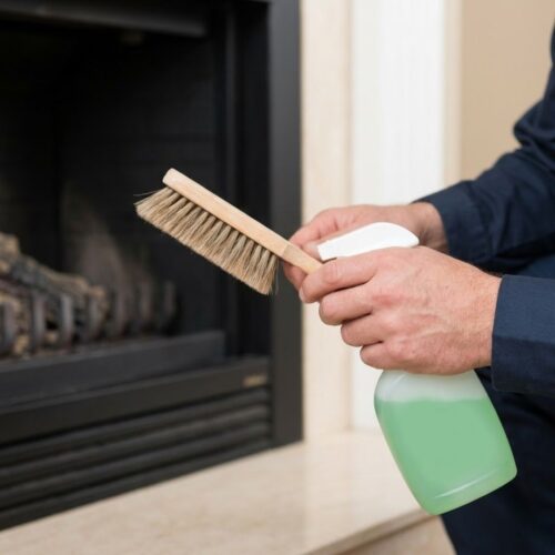 A close-up view shows a professional technician's hands holding a soft brush and non-toxic cleaning spray near a fireplace hearth.